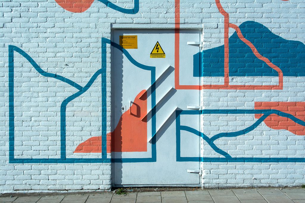 Bright and colorful abstract art featuring bold lines and shapes painted on a white brick wall with a door.