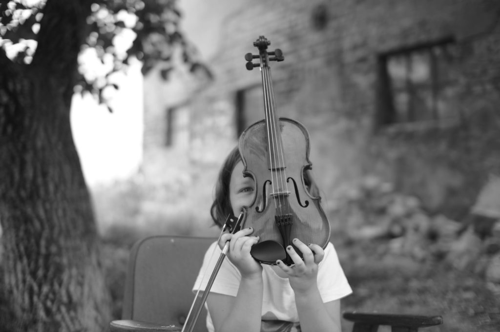 親愛的,如果控制的極致是學會放手? A child holding a violin with a rustic background, captured in black and white for a classic, timeless feel.