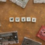 親愛的,如果控制的極致是學會放手? Retro cassette tapes arranged with Scrabble tiles spelling 'Music' on a brown background.