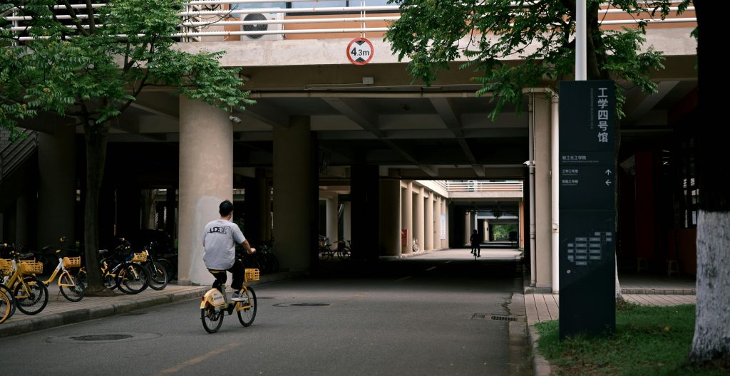 Silhouette of a cyclist riding through a shaded university pathway, surrounded by parked bikes and greenery.