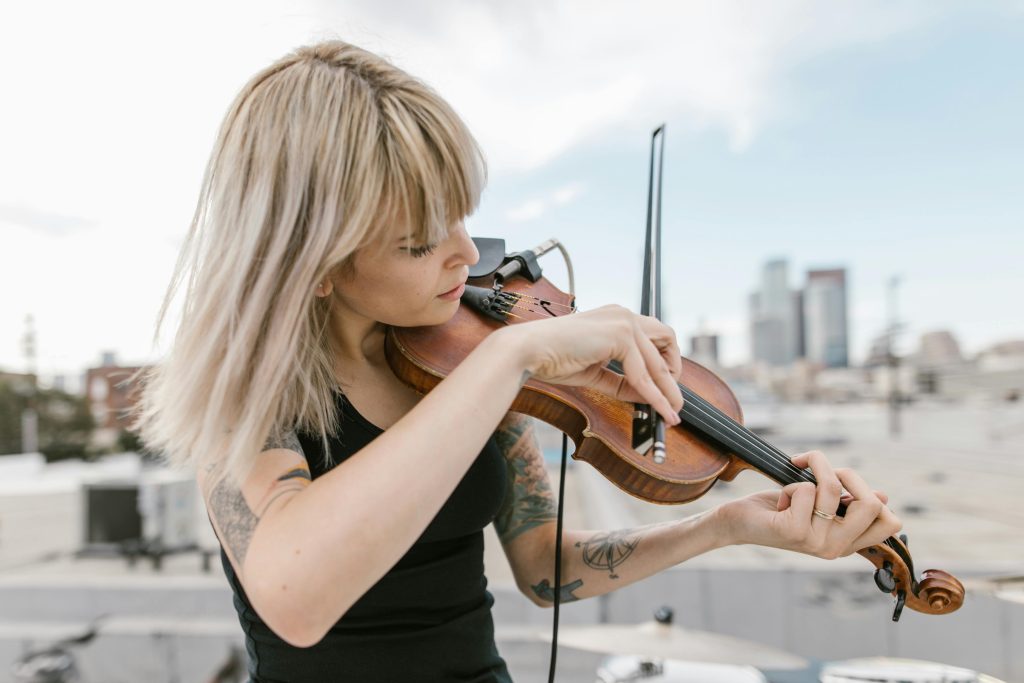 親愛的,如果控制的極致是學會放手? A blonde woman with tattoos plays the violin on a rooftop, city skyline in background.
